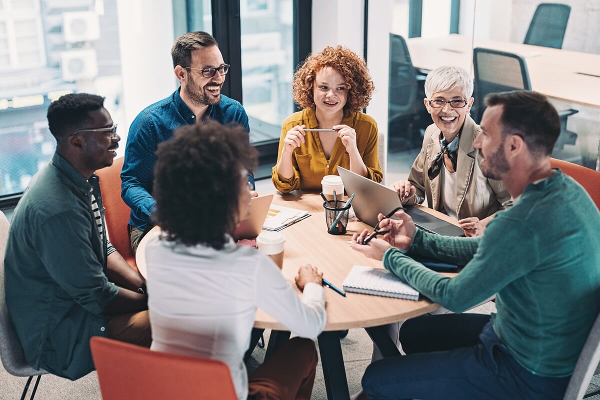 Grupo de pessoas vestindo roupas formais, sentandas ao redor de uma mesa redonda composta por cadernos e notebooks, conversando.