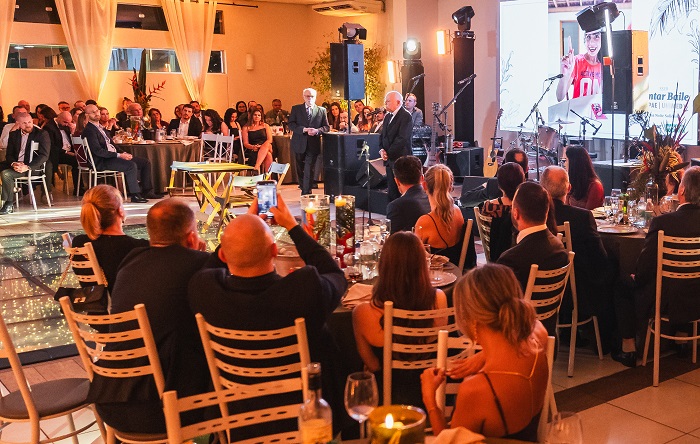 Na primeira imagem, várias pessoas estão sentadas em mesas de um salão elegante, decorado com luzes quentes e cortinas brancas. O público está atento a um evento no palco, onde duas pessoas em trajes formais estão em pé, uma delas segurando um microfone. Ao fundo, há uma grande tela de projeção exibindo uma imagem de uma mulher usando toga e segurando um diploma. Mesas estão decoradas com flores e taças, e algumas pessoas estão de costas, tirando fotos do palco com seus celulares.