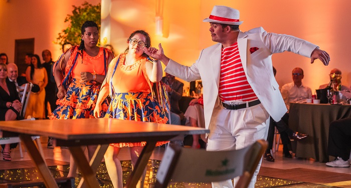 Descrição Imagem: três artistas estão no centro de um salão, performando uma dança. O homem à direita está vestido com calça e paletó brancos, camiseta listrada vermelha e branca, chapéu branco com uma fita vermelha, e está conduzindo as duas mulheres ao seu lado. As duas mulheres estão usando saias coloridas e blusas laranjas. Elas se movimentam enquanto o público, ao fundo, assiste à apresentação. O ambiente é festivo e bem iluminado.