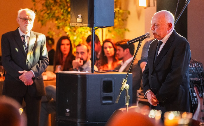 Descrição Imagem: homem de terno preto está em frente a um microfone, fazendo um discurso em um evento formal. Atrás dele, outro homem também de terno, com cabelo grisalho e óculos, observa atentamente. Ao fundo, algumas pessoas estão sentadas em mesas decoradas, enquanto outras prestam atenção no discurso. A iluminação é suave e quente, criando um ambiente acolhedor.