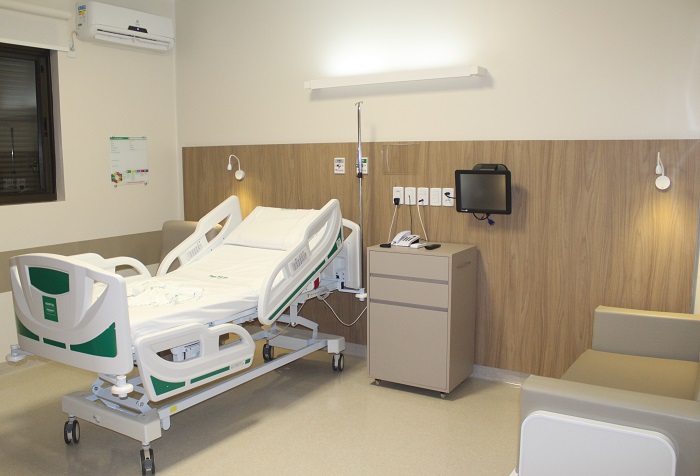 imagem mostra um quarto de hospital moderno e bem equipado, com uma cama hospitalar ajustável, uma poltrona confortável ao lado e uma luminária na parede de madeira clara. Há um painel na parede com tomadas e um monitor, além de uma janela com persianas e um ar-condicionado acima.