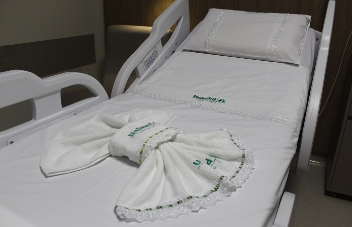 imagem foca na cama hospitalar do mesmo quarto, mostrando detalhes da roupa de cama branca com o logotipo da Unimed. O lençol e o travesseiro estão bem arrumados, e há uma toalha dobrada de forma decorativa sobre a cama.