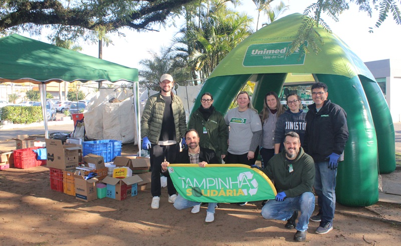 voluntários do projeto tampinha solidaria com voluntários da unimed vale do caí, posam para foto, ao fundo um toldo com a logo da unimed e a frente das pessoas uma bandeira escrita tampinha solidaria